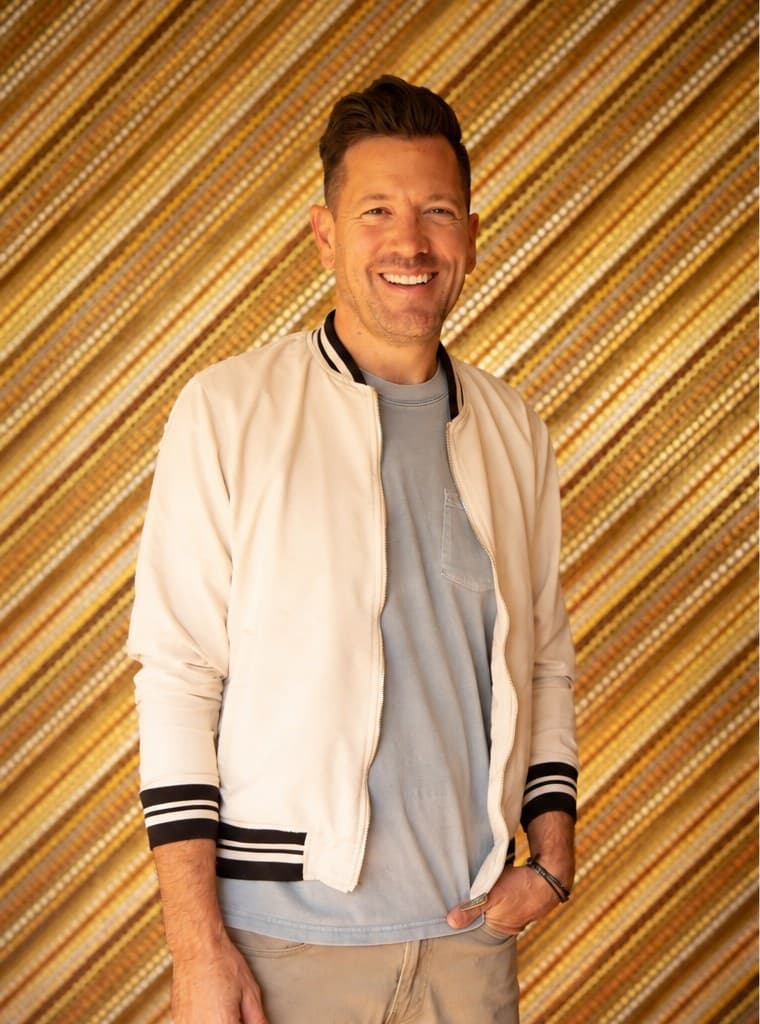 Luke Caldwell smiling in a cream bomber jacket, in front of a gold slatted wall